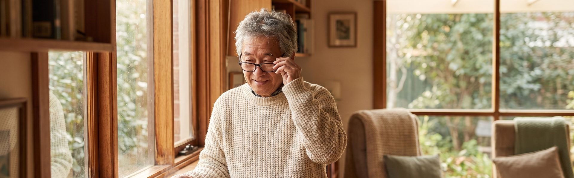 An older man at his sunlit kitchen window in the morning, putting on his reading glasses.